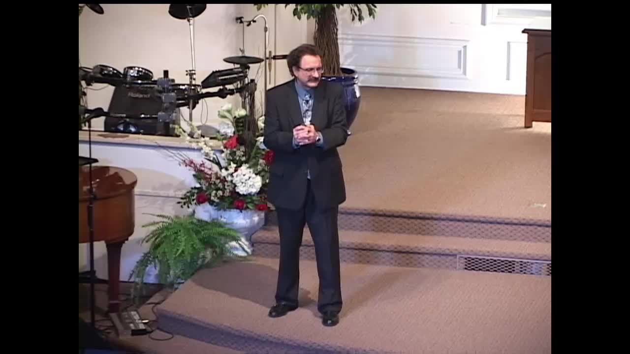 A man in a dark suit stands on a small stage, his hands clasped in front of him. He's facing the camera, likely addressing viewers of the 3ABN Proclaim! Network from the United States.
