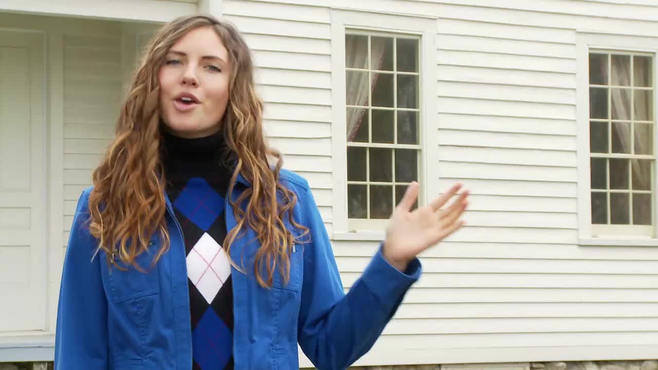 A woman in a blue jacket and argyle sweater is speaking, gesturing with her right hand in front of a white clapboard building. She appears to be on location for a broadcast from the United States, likely for the 3ABN Proclaim! Network.
