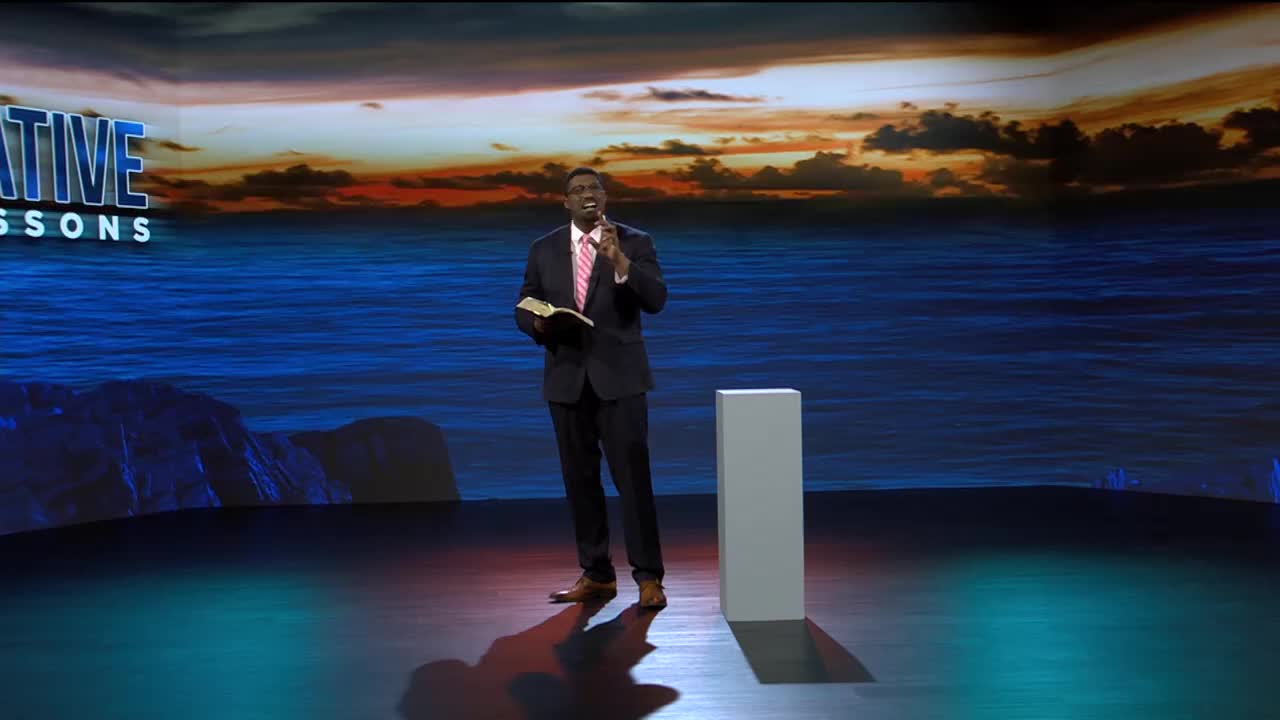 A man in a dark suit, holding a Bible, speaks into a microphone. Behind him, a backdrop depicts a sunset over a calm ocean.
