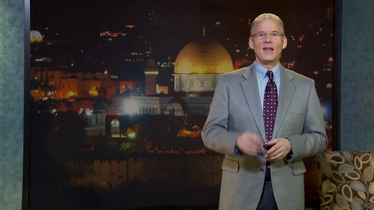 A man in a suit gestures with his hands in front of a backdrop depicting a nighttime cityscape. The golden dome of a building is illuminated, and the scene feels like it's from a television studio in the United States.
