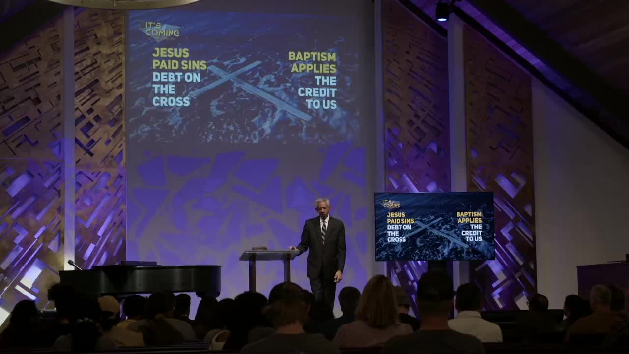 A man in a suit stands at a lectern, gesturing toward a screen displaying text about Jesus and baptism. The audience is seated, facing the stage in what appears to be a church service, likely for the 3ABN Proclaim! Network in the United States.
