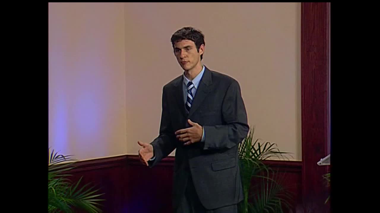 A man in a dark suit gestures with his hands, speaking on a set for 3ABN Proclaim! Network in the United States. His tie has blue and white stripes.
