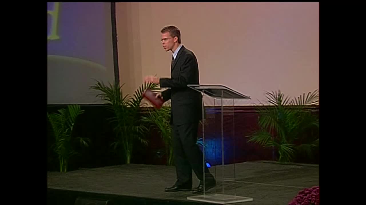 A man in a dark suit, holding a book, gestures as he speaks from behind a clear podium. He's on a stage, likely in the United States, as part of a broadcast for the 3ABN Proclaim! Network.

