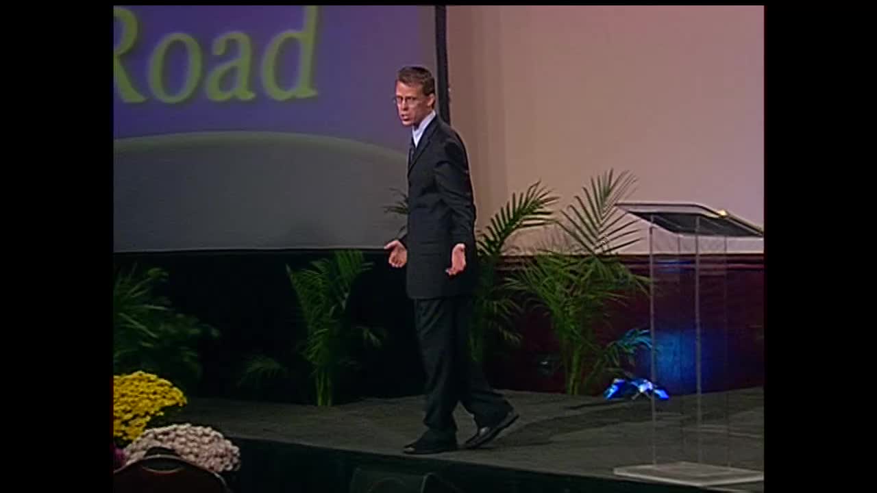 A man in a dark suit strides across a stage, his hands hanging loosely at his sides. Behind him, a screen displays the word "Road" in large, yellow letters.
