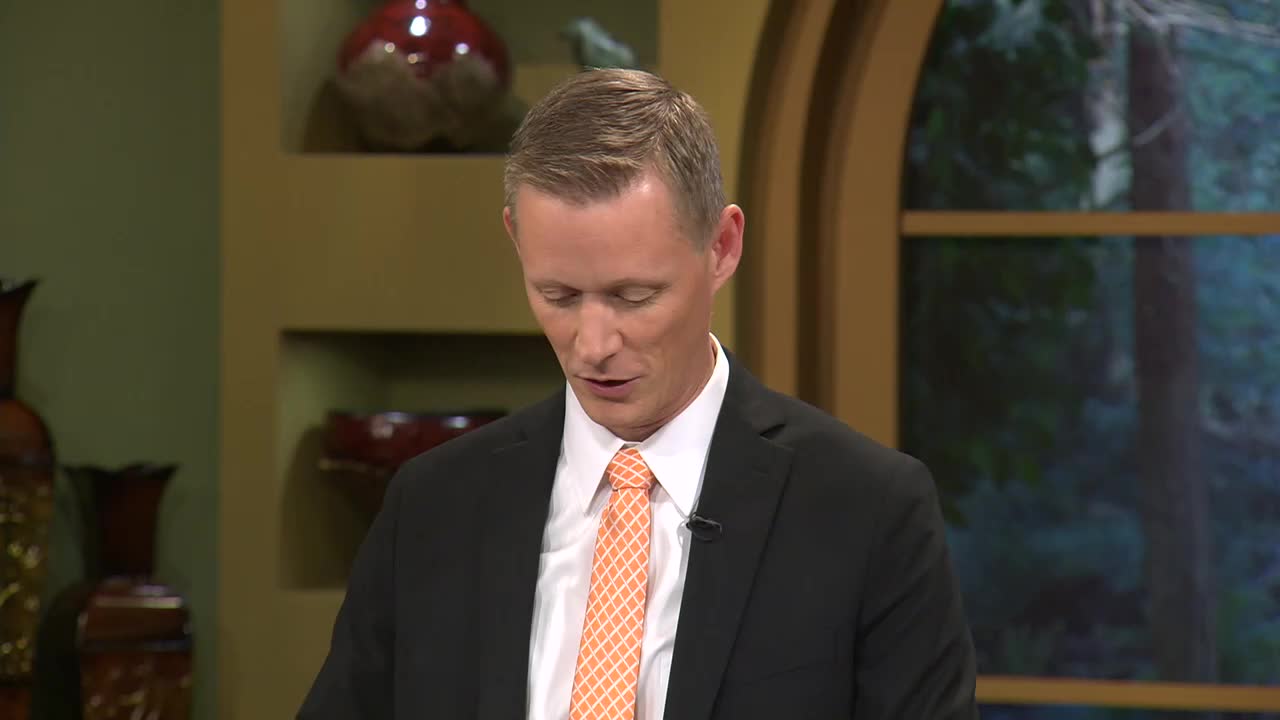 A man in a suit, with a patterned orange tie, is speaking, his eyes focused downward. He's on the set of a program, likely for the 3ABN Proclaim! Network in the United States.
