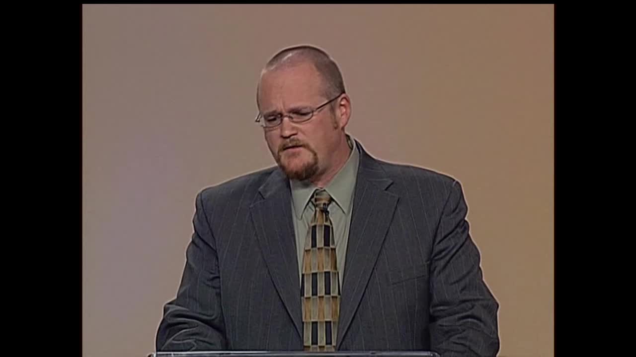 A man in a suit, speaking on the 3ABN Proclaim! Network, is looking down as if reading or reflecting. His tie has a geometric pattern, and he's wearing glasses.
