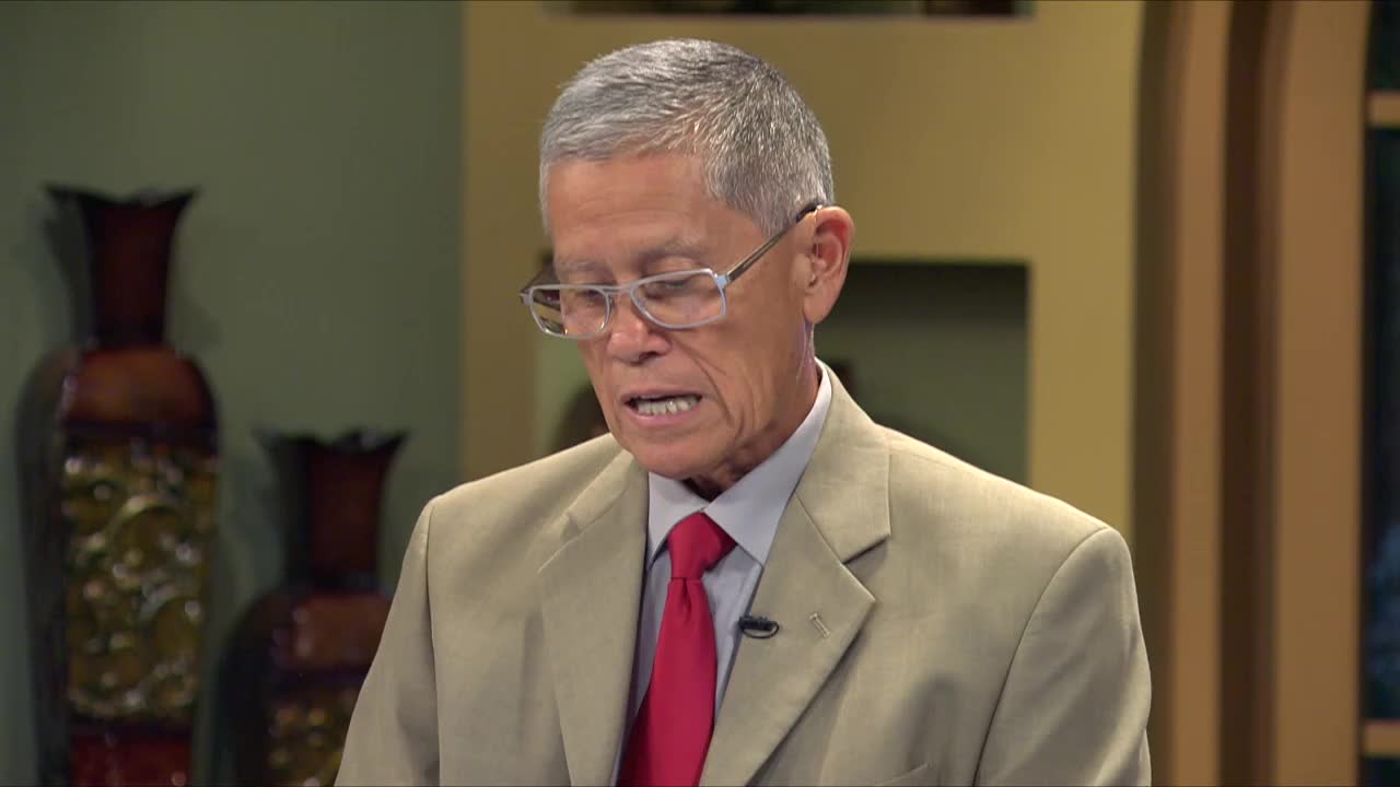 A man in a suit and tie, likely on 3ABN Proclaim! Network from the United States, is speaking, his mouth moving as he reads. He's wearing glasses and a red tie, and the background includes decorative vases.

