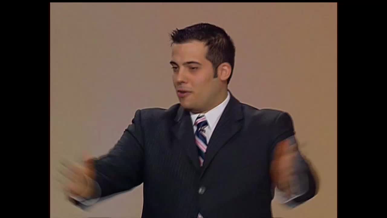 A man in a suit gestures expansively, his hands a blur as he speaks. He's on a set, likely for a broadcast on the 3ABN Proclaim! Network, in the United States.

