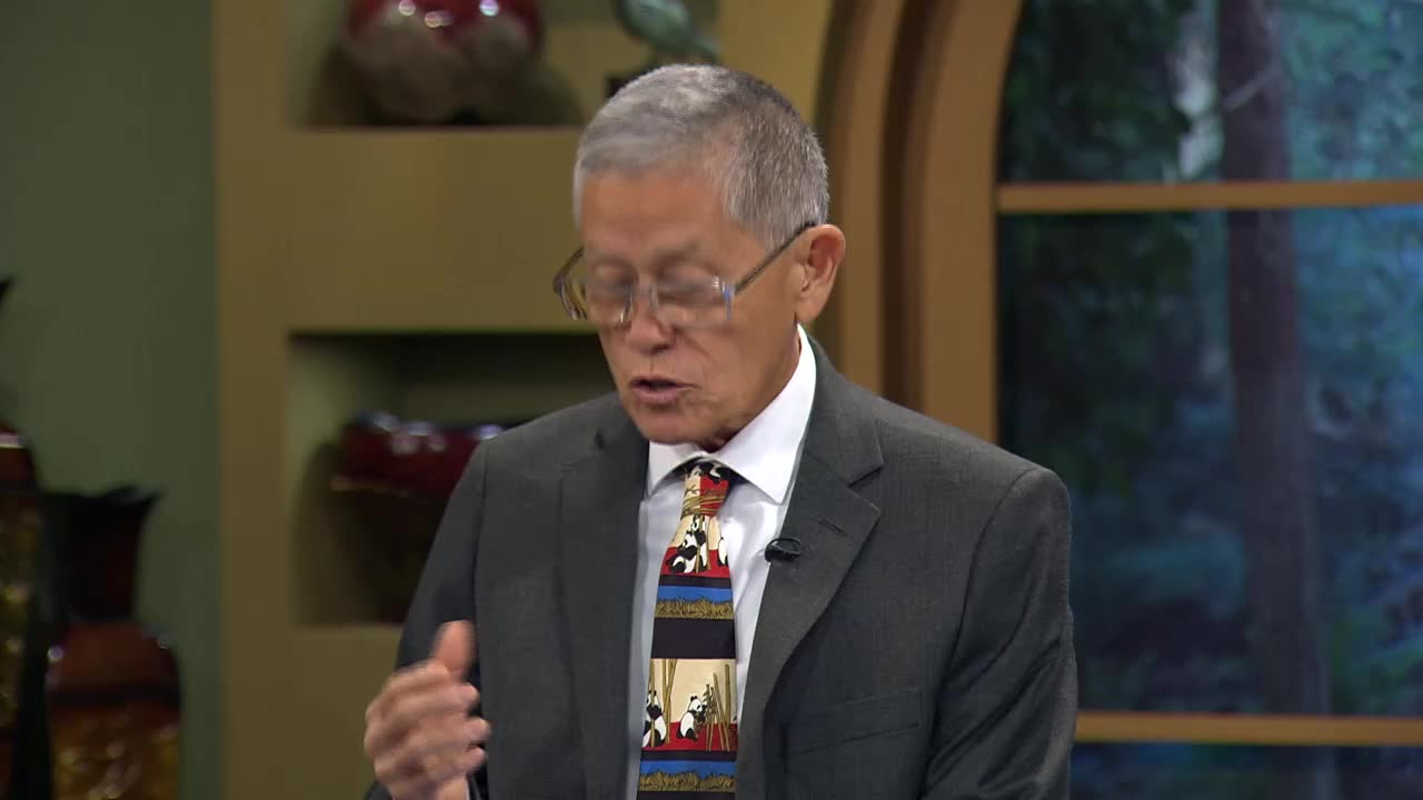 A man in a suit, wearing glasses and a patterned tie, speaks into a microphone. His hands are moving as he gestures, likely on the set of a 3ABN Proclaim! Network program in the United States.
