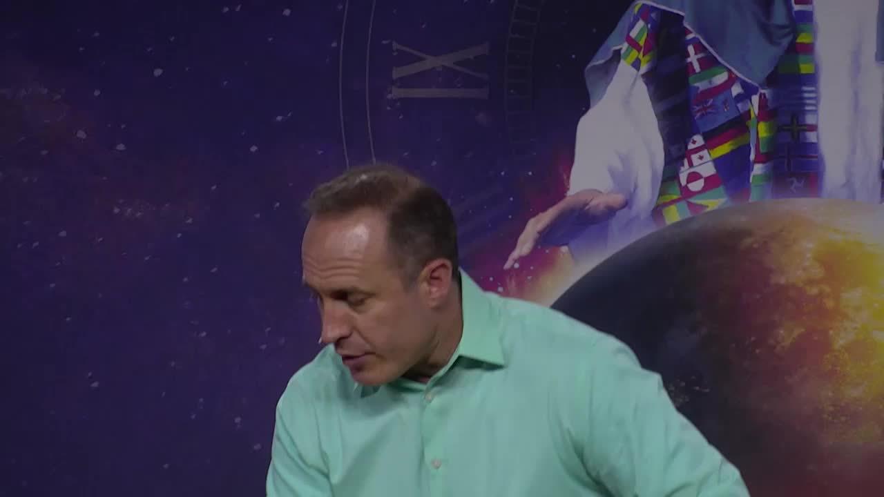 A man in a light green shirt leans forward, speaking. Behind him, a celestial backdrop includes a clock face and a globe.
