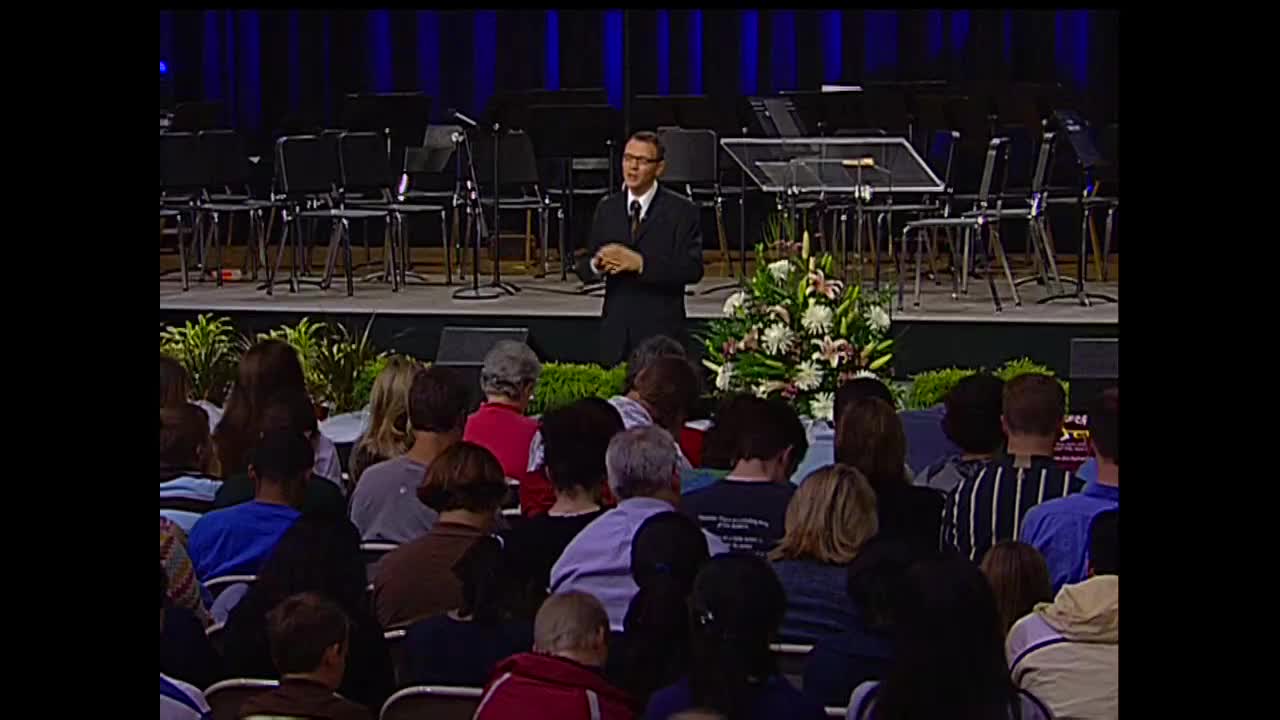 A man in a suit gestures as he speaks, standing on a stage decorated with flowers and a backdrop of empty chairs. The audience is seated, facing the stage, watching the presentation.
