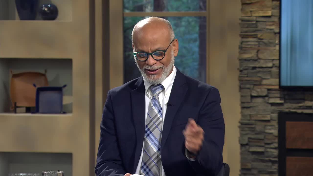 A man in a navy suit and glasses gestures with his right hand as he speaks. He's on the set of a program, likely for the 3ABN Proclaim! Network in the United States.
