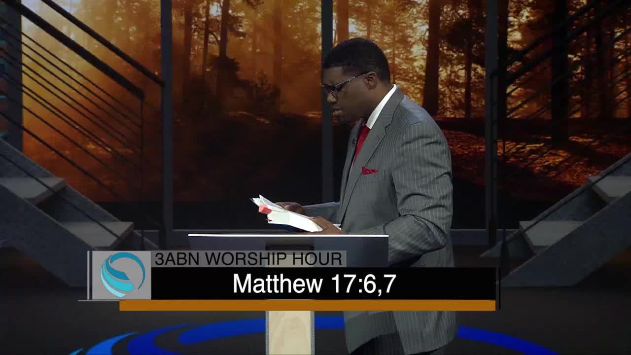 A man in a gray suit stands behind a podium, reading from a book. The screen below him displays the 3ABN Worship Hour logo and a Bible verse.

