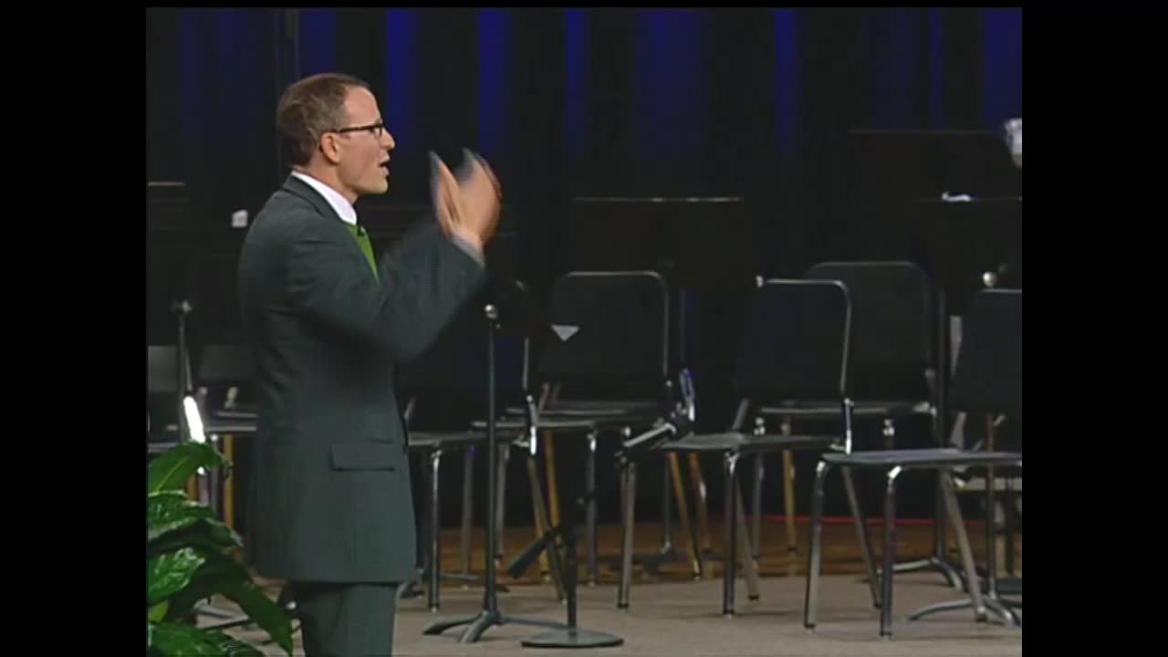 A man in a green suit gestures with his hands, speaking into a microphone. Behind him, rows of empty chairs await the musicians for a 3ABN Proclaim! Network broadcast in the United States.
