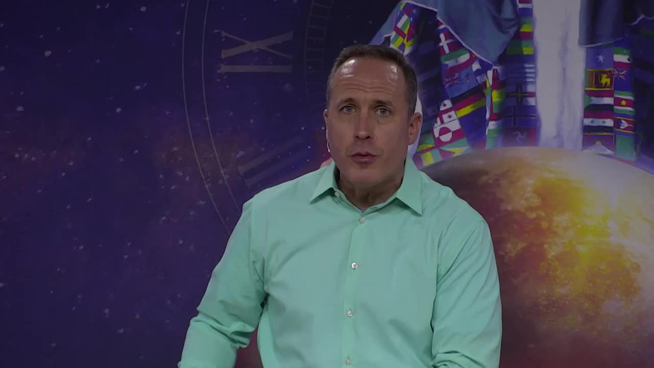 A man in a light green shirt speaks directly to the camera, his expression serious. Behind him, a backdrop features a globe and what looks like flags representing different nations.
