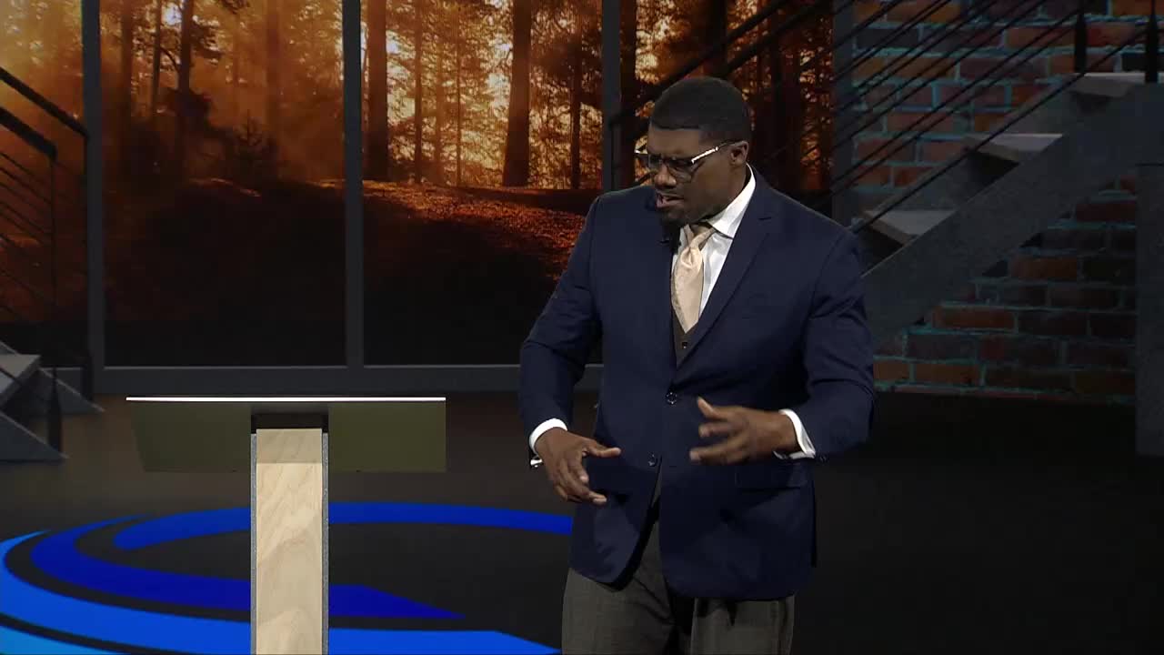 A man in a navy suit gestures with his hands near a podium, speaking in front of a backdrop of a forest scene. The set looks like it's from a broadcast on the 3ABN Proclaim! Network in the United States.
