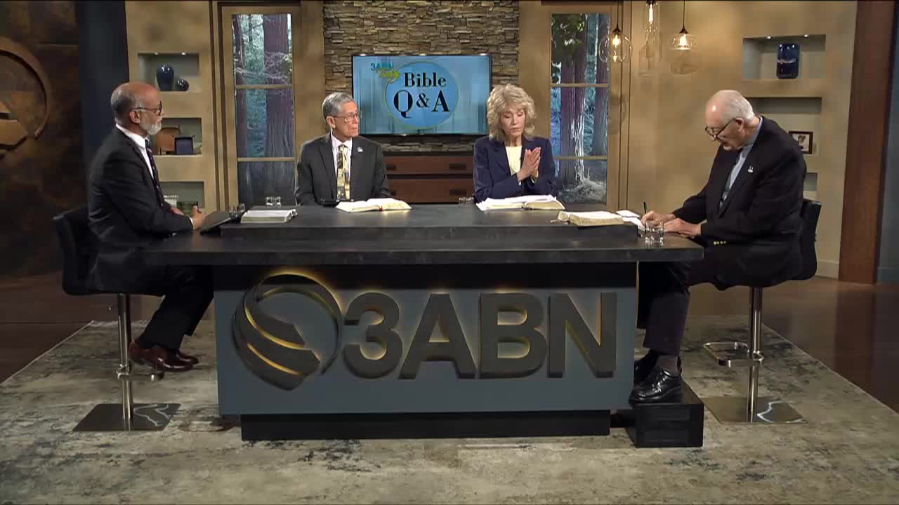 Four people sit around a desk, a "Bible Q&A" logo on the monitor behind them. The woman in the middle claps her hands, while the man on the right leans forward, writing on a notepad.
