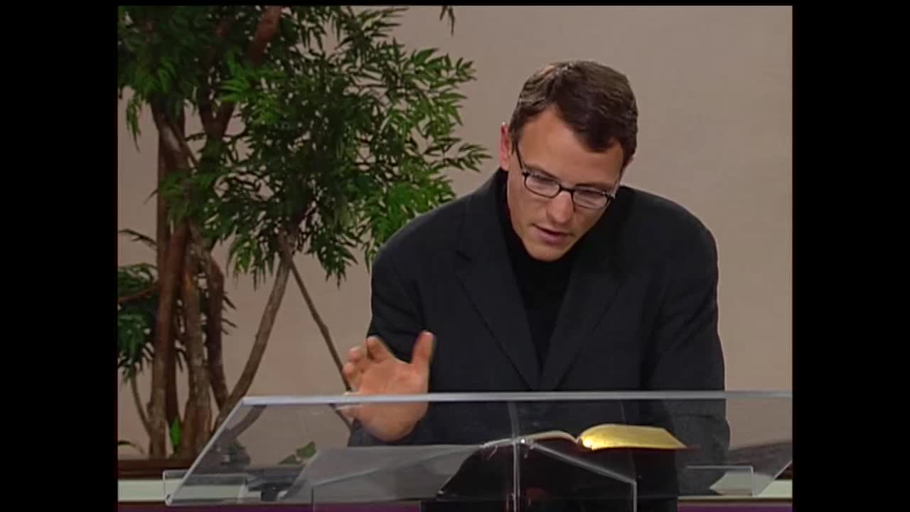 A man in a black suit jacket and turtleneck leans forward, gesturing with his left hand as he speaks. He looks down at an open book with gold-edged pages resting on a clear lectern.