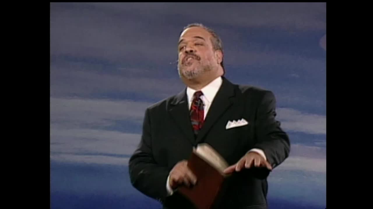 The speaker, dressed in a dark suit and red tie, gestures with his right hand while holding a book. He's on a stage with a blue, cloudy backdrop, likely for a broadcast on the 3ABN Proclaim! Network in the United States.