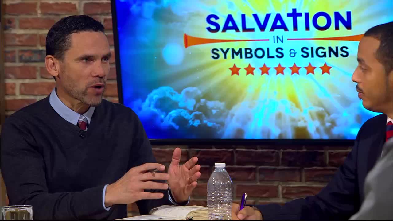 A man in a dark sweater gestures with his hands as he speaks, while a screen behind him displays "SALVATION IN SYMBOLS & SIGNS." Another man in a suit sits to his right, listening intently.