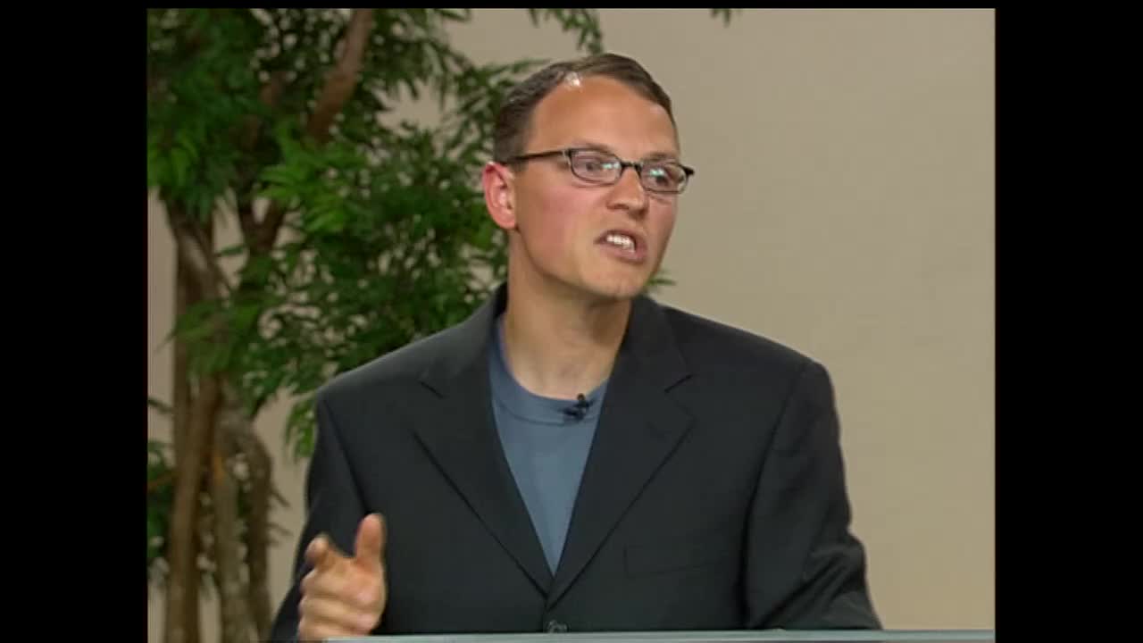 A man in a dark suit and glasses speaks animatedly, gesturing with his left hand. He's on a set, with a leafy plant behind him, likely for a broadcast on the 3ABN Proclaim! Network.