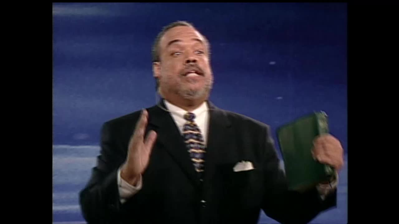 A man in a suit gestures with his left hand while holding a green book in his right. He speaks with conviction, his expression animated against a blue backdrop.