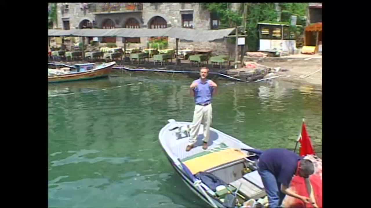 A man in a blue shirt and tan pants stands on a small boat, gesturing with his hands. Another man, wearing a dark blue shirt and jeans, kneels in the back of the boat, tending to equipment.