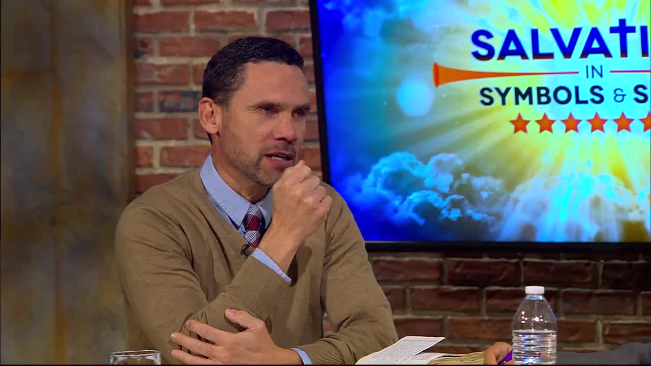 A man in a tan sweater and collared shirt leans forward, resting his chin on his hand. Behind him, a screen displays "Salvation in Symbols & Signs" with a bright, sunburst graphic.