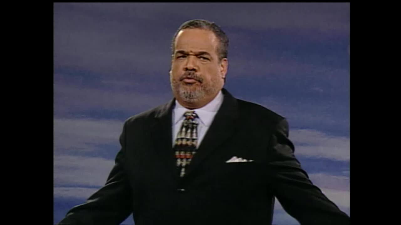 A man in a suit stands with his arms outstretched, his gaze fixed forward. He speaks with conviction, his voice resonating through the studio.