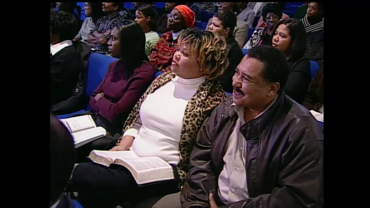 A man with a mustache smiles and speaks, his leather jacket open over a collared shirt. Beside him, a woman in a leopard-print jacket leans forward, her gaze directed upward.