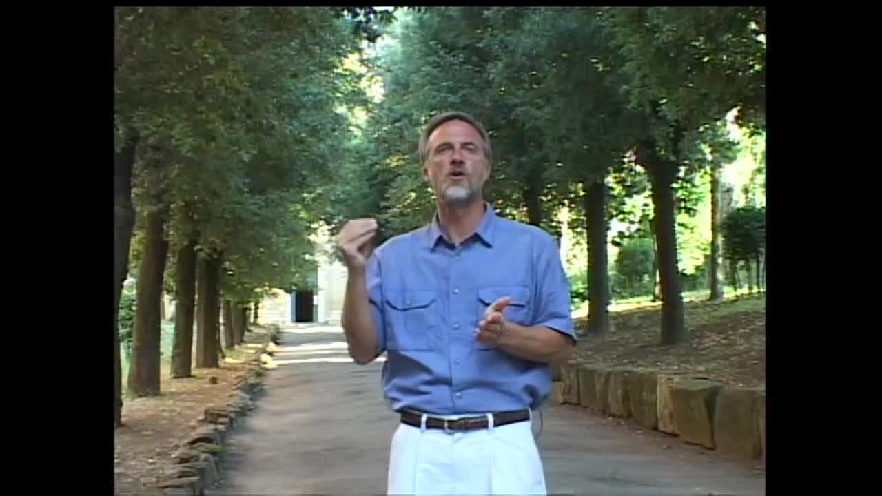A man in a blue shirt and white pants stands on a paved path, gesturing with his hands as he speaks. Tall trees line the path on both sides, creating a shaded, natural setting.