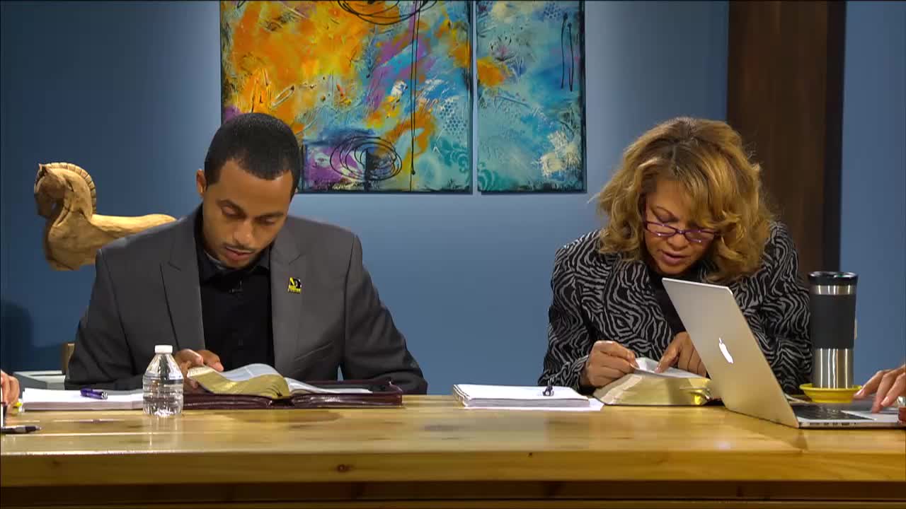 A man in a grey suit leans over a large open book, his finger tracing a line of text. Across from him, a woman in a patterned jacket also studies a book, a laptop open beside her.