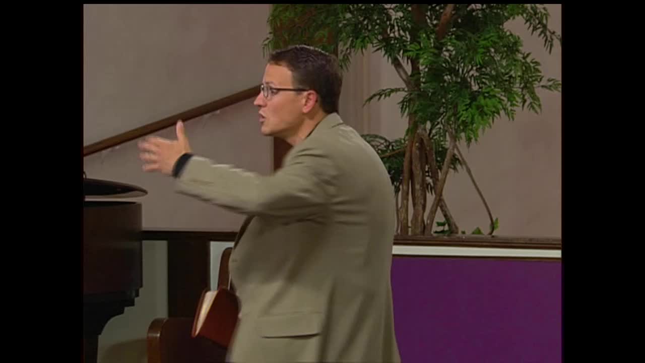 A man in a tan suit gestures with his left hand while speaking, his glasses reflecting the light. He stands near a piano in what appears to be a church setting, with a potted plant to his right.