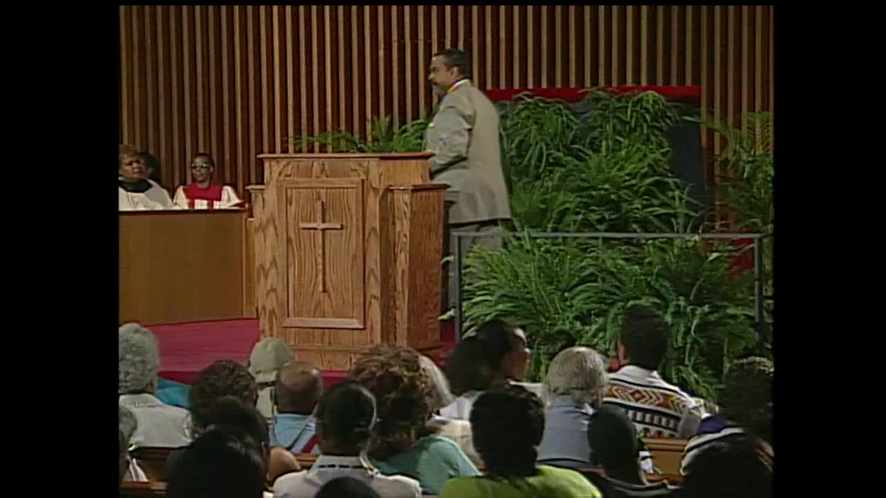 A man in a light suit walks away from a wooden pulpit. Lush green plants fill the background behind him.