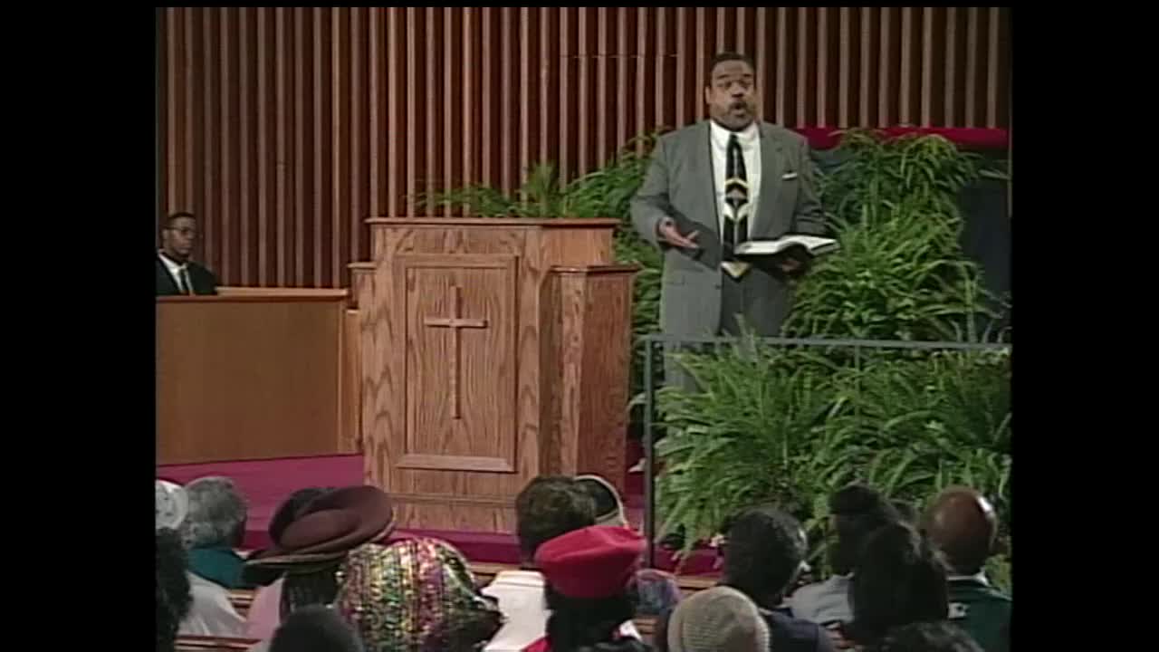 A preacher in a grey suit stands at a wooden pulpit, holding an open Bible. He gestures with his left hand while speaking to the congregation gathered in front of him.