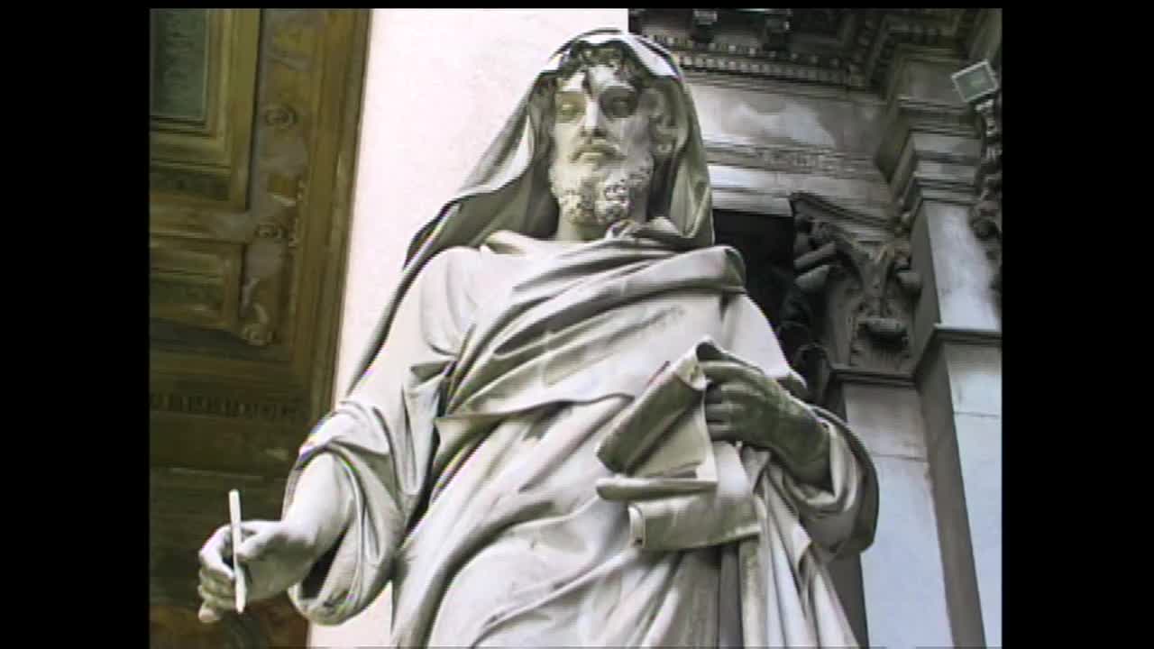 A stone figure, draped in flowing robes, holds a stylus and a scroll. The weathered stone suggests it has stood for a long time.
