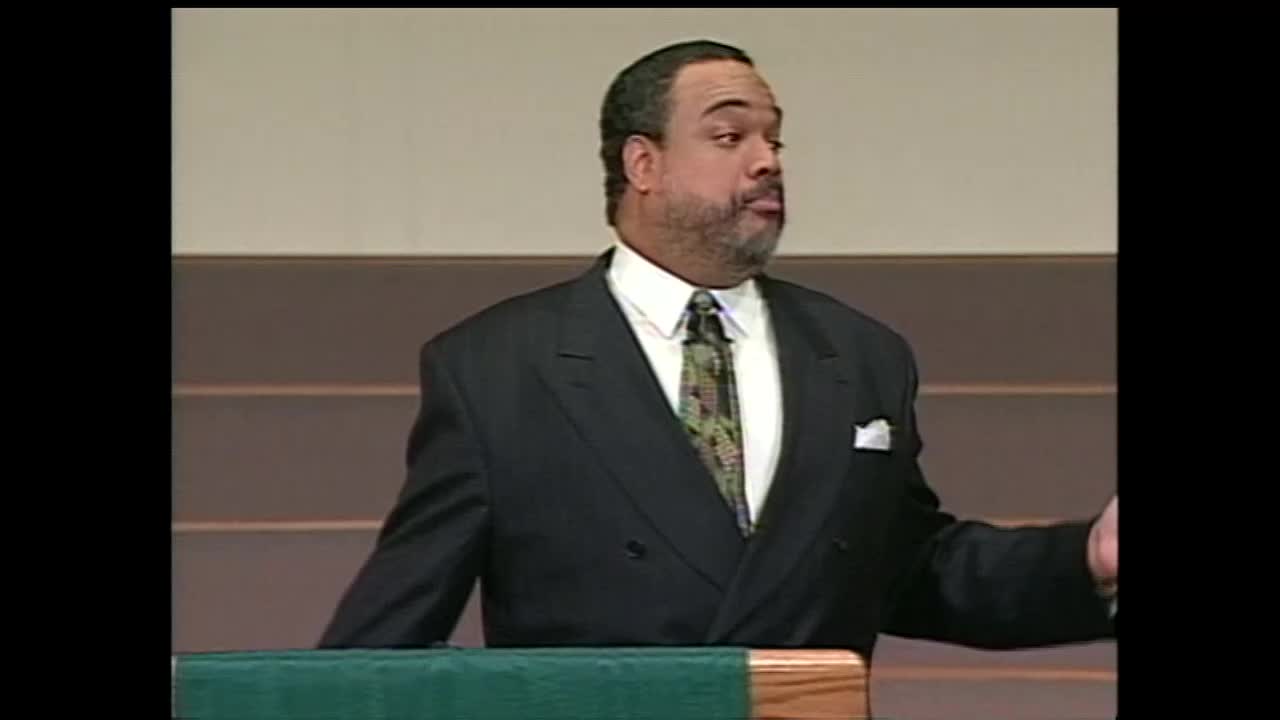 A man in a dark suit and patterned tie gestures with his right hand as he speaks. He stands behind a wooden podium covered with a green cloth.