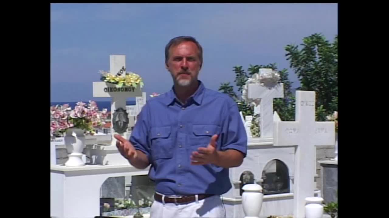 A man in a blue shirt stands in a cemetery, gesturing with his hands. White crosses and floral arrangements fill the background under a bright sky.