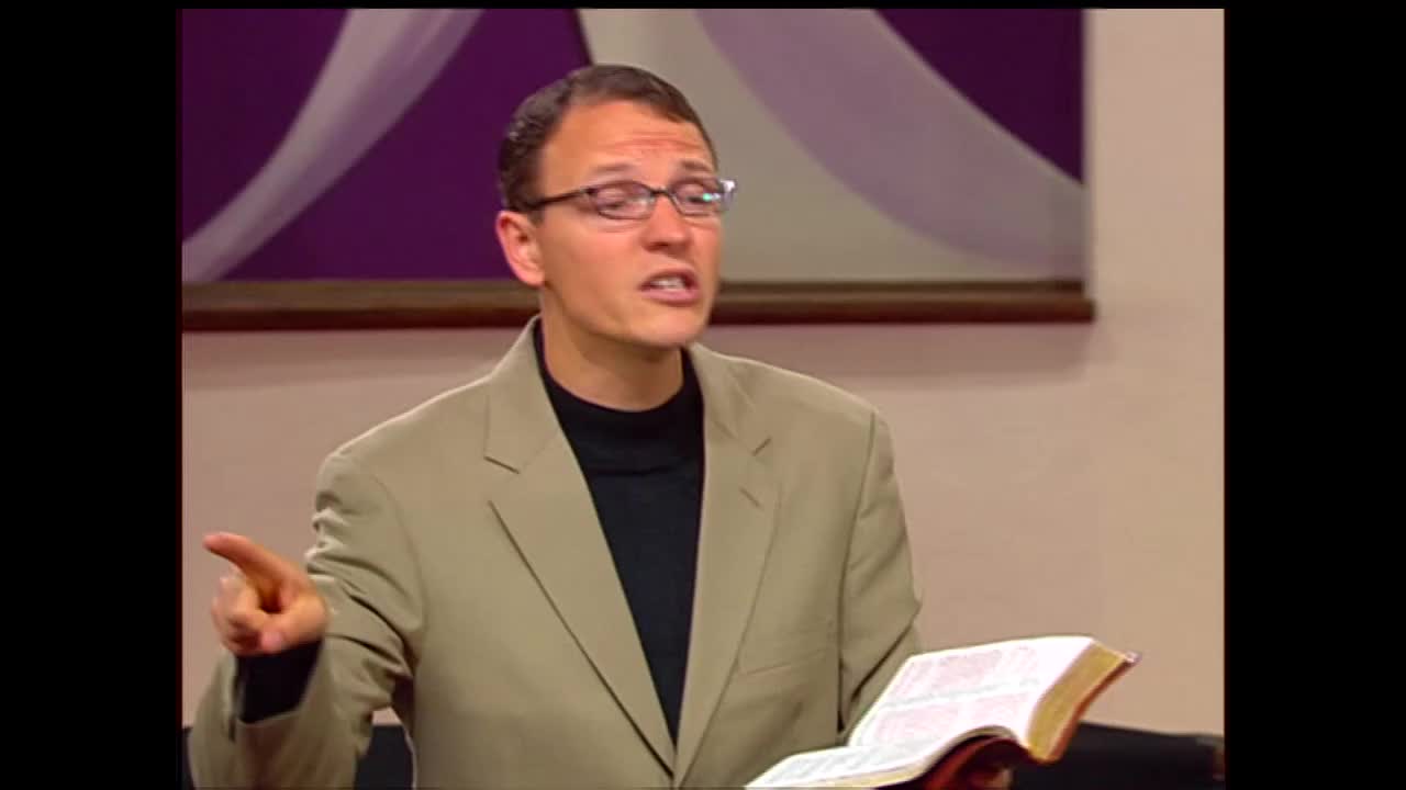 A man in a tan suit jacket and black turtleneck gestures with his left hand while holding an open Bible. He speaks with conviction, his glasses reflecting the studio lights.