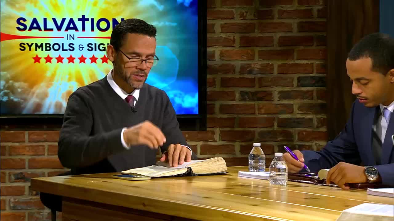 A man in a dark sweater gestures with his hand over an open Bible. To his right, another man in a suit writes with a purple pen. Behind them, a screen displays "SALVATION IN SYMBOLS & SIGNS" with a burst of light.