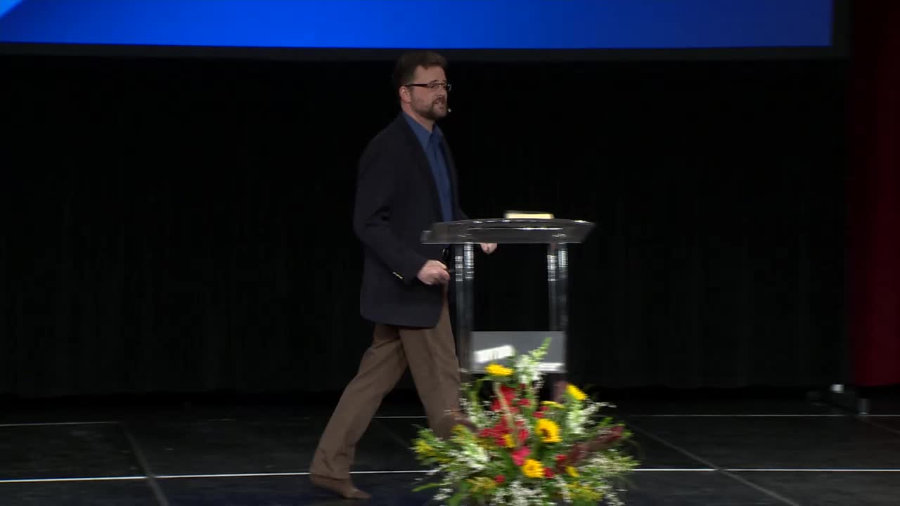A man in a dark jacket and brown pants walks across a stage, his left hand reaching towards a clear podium. A vibrant floral arrangement sits at the edge of the stage, just past the podium.