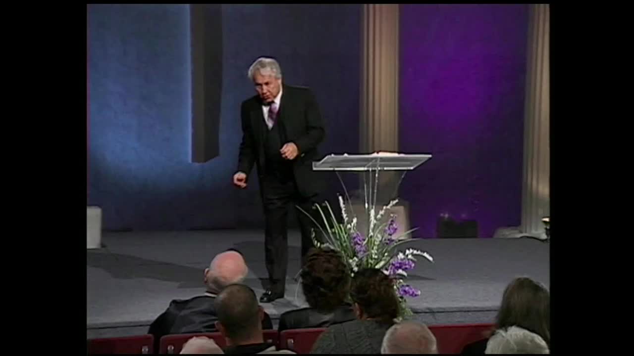 A man in a suit gestures as he speaks from behind a podium. The 3ABN Proclaim! Network broadcast shows a seated audience before him.