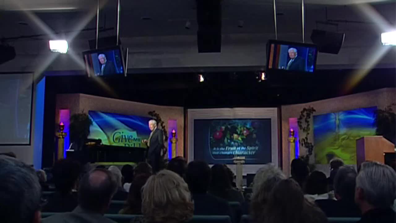 A man in a dark suit stands on a stage, gesturing as he speaks. A screen behind him displays "Give to the Lord," and another shows a colorful still life of fruit. The audience watches attentively.