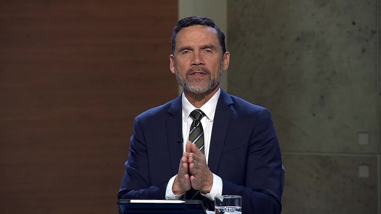 A man in a dark suit and tie clasps his hands together, speaking directly to the camera. A glass with the 3ABN Proclaim! Network logo sits on the table before him.