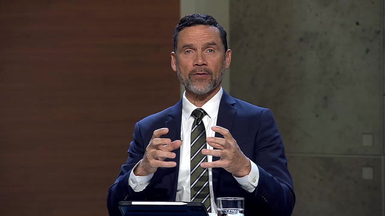 A man in a suit and tie gestures with his hands as he speaks. He's seated at a table, with a tablet and a glass of water in front of him, likely during a broadcast for the 3ABN Proclaim! Network.