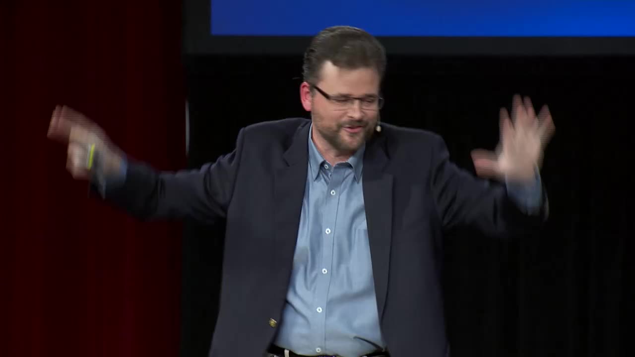 A man in a dark suit jacket and blue shirt gestures with both hands, his right arm extended and his left arm raised. He speaks animatedly on a stage, likely during a presentation for the 3ABN Proclaim! Network in the United States.