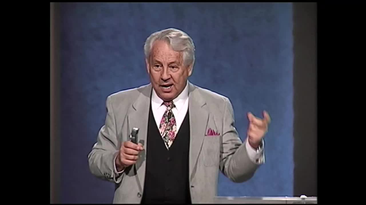 A man in a light gray suit, black vest, and floral tie gestures with his right hand as he speaks. He holds a silver object in his left hand, standing before a blue backdrop. This moment is part of a broadcast on the 3ABN Proclaim! Network.
