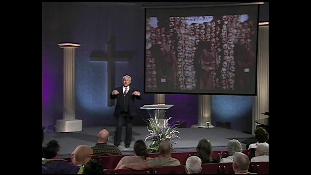 A man in a suit gestures with his hands while speaking on a stage, a large projection behind him displays a wall of skulls. The audience watches attentively from their seats.