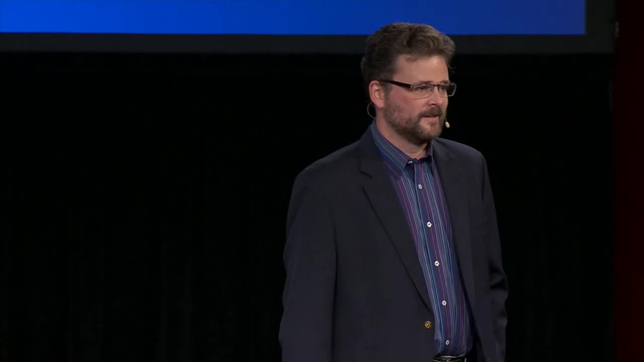 A man with glasses and a beard stands on a stage, speaking into a microphone. He wears a dark suit jacket over a striped shirt, likely for a presentation on the 3ABN Proclaim! Network.