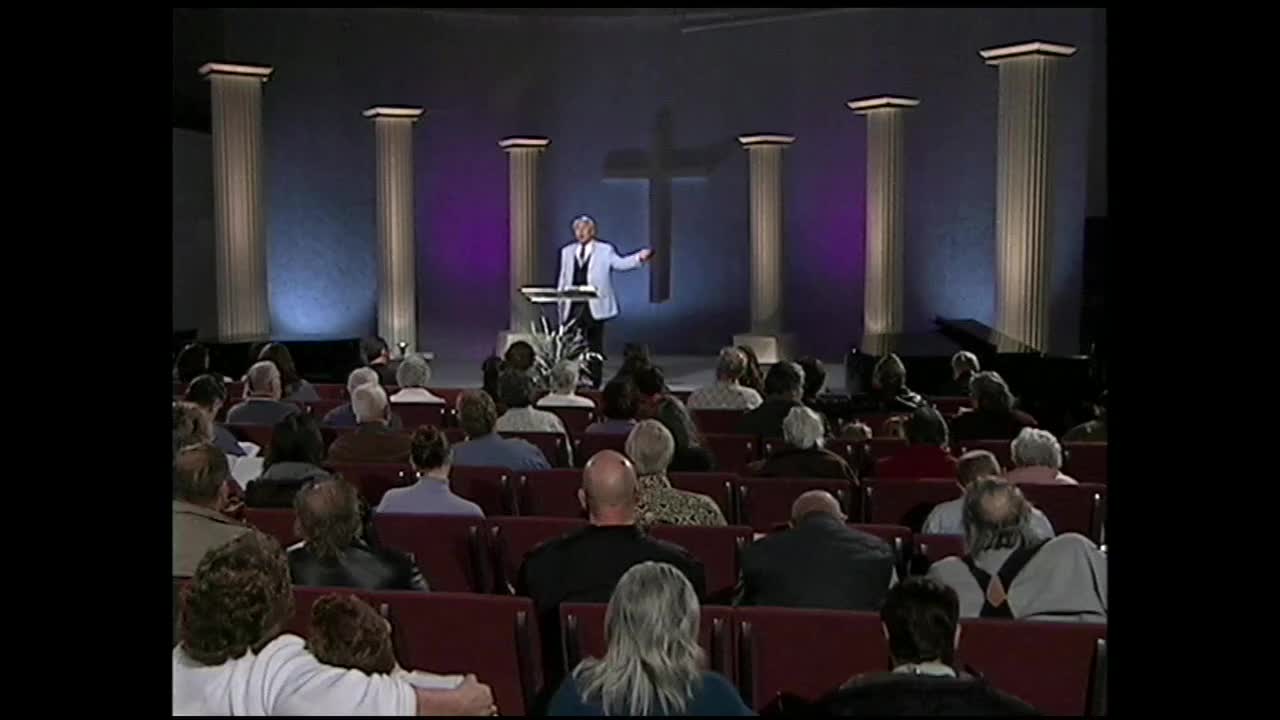 A man in a suit stands at a podium, gesturing to the audience. Rows of people fill the seats before him, their faces turned towards the stage.